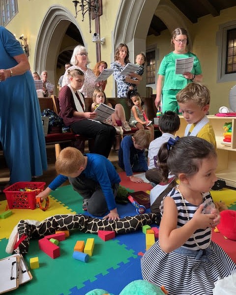 children in the foreground using soft toys to engage in worship while parents and staff in background offer supervision