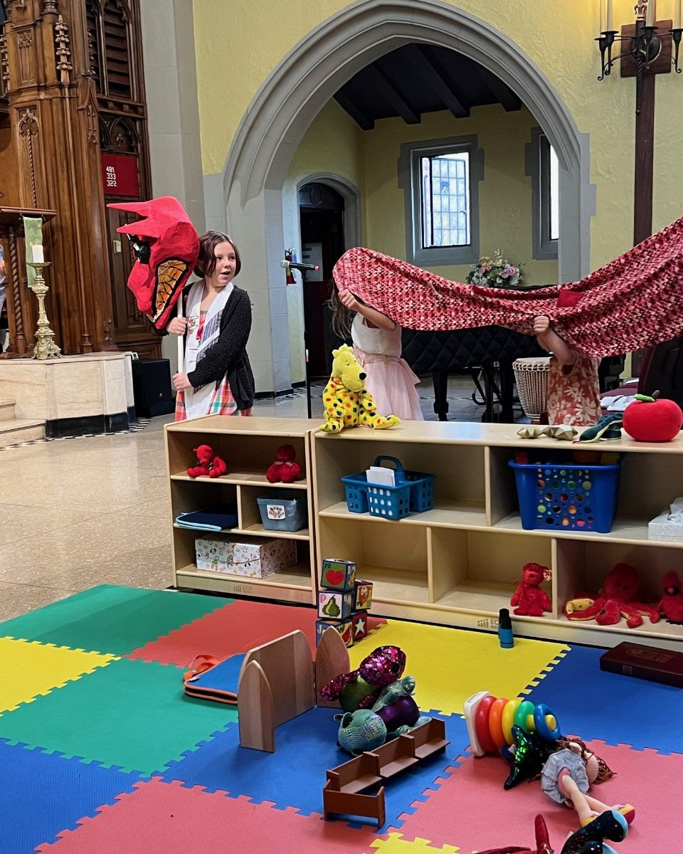 toys in a colorful Kid's Corner in the foreground, and children processing down the aisle with a large dragon costume in the background